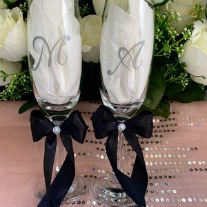 May include: Two champagne flutes with black satin bows and rhinestones. The flutes are personalized with the letters "M" and "A" in silver.