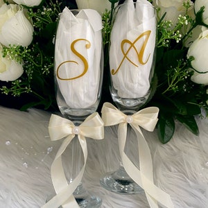 May include: Two champagne flutes with gold lettering, "S" and "A", tied with ivory satin bows and pearl accents. The flutes are on a white fluffy surface.