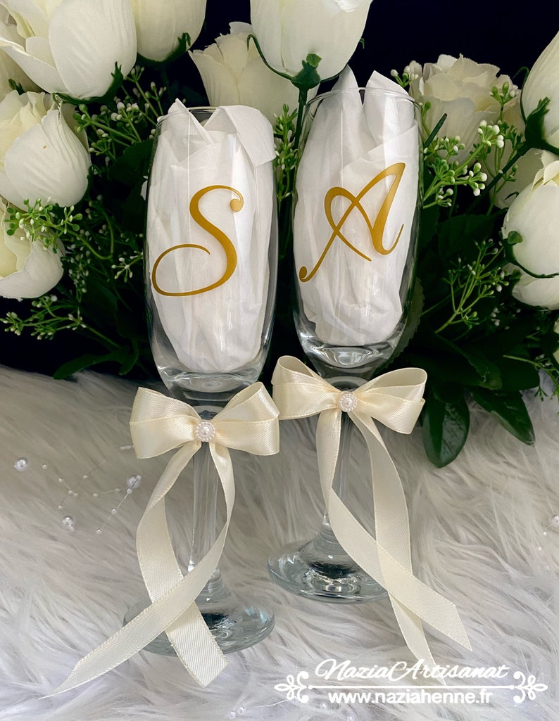 May include: Two champagne flutes with gold lettering, "S" and "A", tied with ivory satin bows and pearl accents. The flutes are on a white fluffy surface.