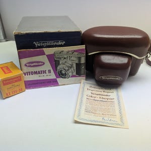 May include: A vintage camera kit featuring a brown leather camera case, a Voigtländer Vitomatic II camera box, a Kodak film box, and an instruction manual. The kit includes a camera and accessories.