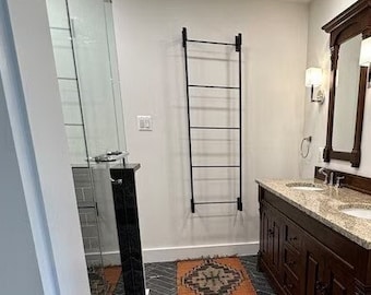 May include: A bathroom with a double vanity with a granite countertop and dark wood cabinets. The vanity has two sinks and two mirrors. The bathroom has a black and white tiled floor with a patterned rug. There is a towel rack on the wall.