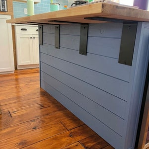 May include: A close-up of a kitchen island with a light wood countertop and a blue painted base. The island is supported by three black metal brackets. The island is in a room with light wood floors.