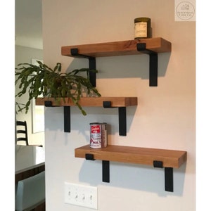 May include: Three wooden shelves with black metal brackets mounted on a white wall. The shelves are empty except for a small candle on the top shelf and a can of food on the middle shelf.