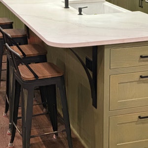 May include: A white kitchen island with a curved countertop and a farmhouse style sink. The island has a black metal bracket supporting the countertop and features green cabinets with drawers. Three black metal bar stools with wooden seats are positioned in front of the island.