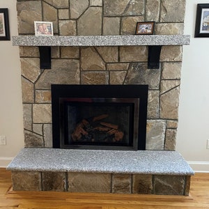 May include: A stone fireplace with a black metal surround and a granite hearth. The fireplace has a wood-burning insert with logs inside. There are two small framed pictures on the mantel above the fireplace.