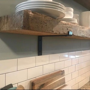 May include: A rustic wooden shelf with a rough-hewn texture, supported by black metal brackets. The shelf displays white ceramic plates, bowls, and cups. The shelf is mounted on a light gray wall above white subway tiles.