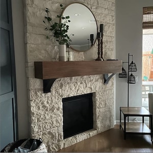 May include: A white stone fireplace with a wooden mantel and a black fireplace insert. A round gold mirror hangs above the mantel with a green plant in a white vase. A black metal floor lamp with three woven shades stands to the right of the fireplace.