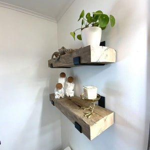 May include: Two wooden shelves with black metal brackets mounted on a white wall. The top shelf has a potted plant with green leaves and a white pot. The bottom shelf has a candle on a gold bird-shaped stand and three glass jars with cork lids.