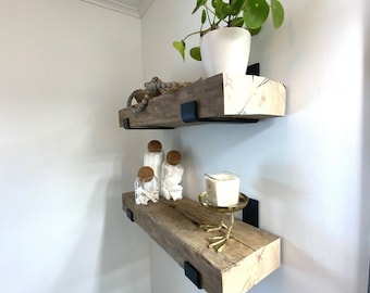 May include: Two wooden shelves with black metal brackets mounted on a white wall. The top shelf has a potted plant with green leaves and a white pot. The bottom shelf has a candle on a gold bird-shaped stand and three glass jars with cork lids.