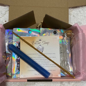 May include: A pink and white gift box with a variety of items for better sleep. The box contains a blue candle, a small bottle of clear crystals, a stick of incense, and a card with the words "Better Sleep" and a list of ingredients.