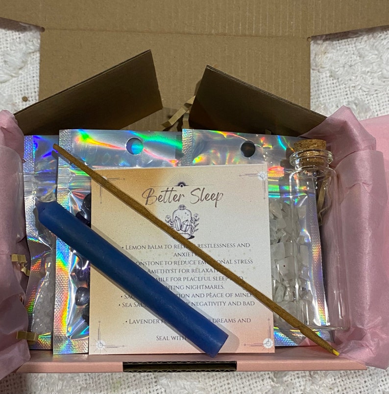 May include: A pink and white gift box with a variety of items for better sleep. The box contains a blue candle, a small bottle of clear crystals, a stick of incense, and a card with the words "Better Sleep" and a list of ingredients.