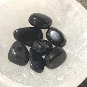 May include: A collection of seven smooth, black obsidian tumbled stones in a white bowl. The stones are various sizes and shapes, with a glossy finish.