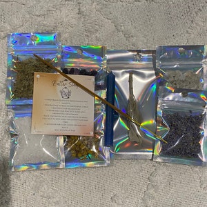 May include: A Better Sleep spell kit with a blue candle, sage, lavender, moonstone, amethyst, chamomile, lemon balm, sea salt, and a wooden stick of incense. The kit is packaged in small clear plastic bags with a holographic iridescent finish. The text on the card reads: "Better Sleep - Lemon balm to relieve restlessness and anxiety - Moonstone to reduce emotional stress - Amethyst for relaxation - Chamomile for peaceful sleep and preventing nightmares - Sage for protection and peace of mind - Sea salt to keep away negativity and bad vibes - Lavender for sleep, good dreams and peace - Seal with blue wax"