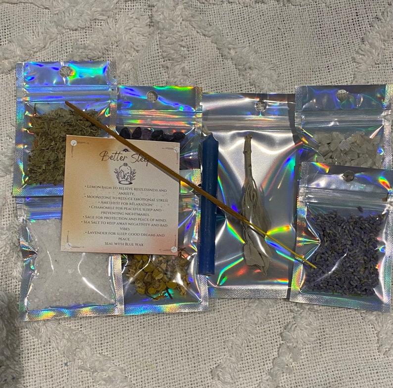 May include: A Better Sleep spell kit with a blue candle, sage, lavender, moonstone, amethyst, chamomile, lemon balm, sea salt, and a wooden stick of incense. The kit is packaged in small clear plastic bags with a holographic iridescent finish. The text on the card reads: "Better Sleep - Lemon balm to relieve restlessness and anxiety - Moonstone to reduce emotional stress - Amethyst for relaxation - Chamomile for peaceful sleep and preventing nightmares - Sage for protection and peace of mind - Sea salt to keep away negativity and bad vibes - Lavender for sleep, good dreams and peace - Seal with blue wax"