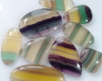 May include: A collection of polished fluorite gemstones in various shapes, including oval, teardrop, and irregular forms. The stones display bands of color, with hues of yellow, purple, green, and cream. The gemstones are arranged on a white surface.