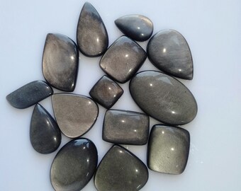 Könnte beinhalten: Eine Sammlung polierter Obsidian-Edelsteine in verschiedenen Formen, darunter Tropfen, Oval und Rechteck. Die Steine haben eine dunkle, reflektierende Oberfläche mit metallischem Glanz. Die Edelsteine sind auf einer weißen Oberfläche angeordnet.