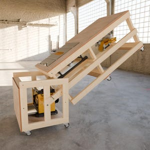 May include: A light-colored wooden workbench on casters featuring a hinged, tilting top surface. A yellow and black power tool is mounted within the lower frame, while a second yellow and black power tool is attached to the underside of the tilting top.