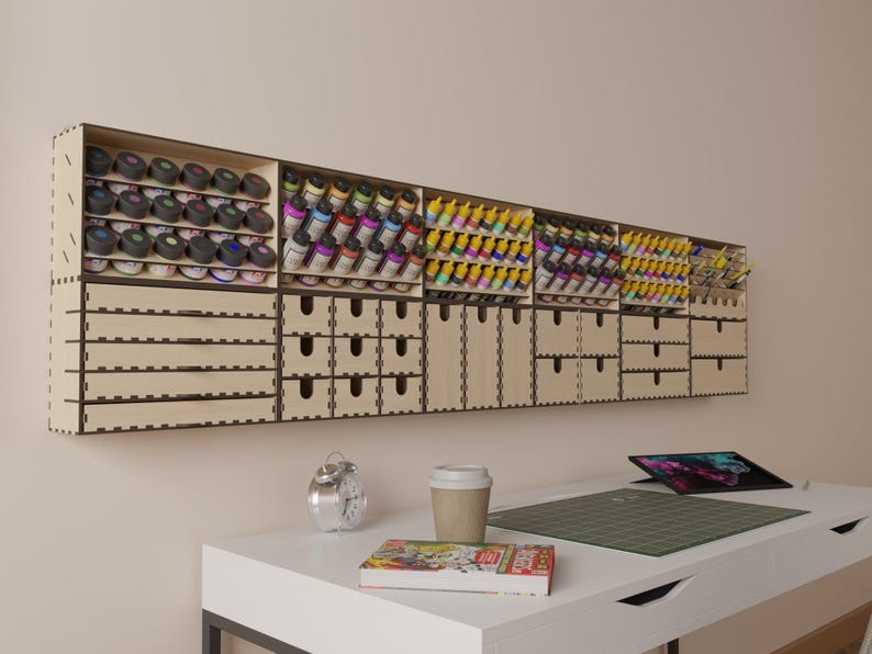 May include: A wall-mounted wooden storage unit with multiple drawers and shelves for organizing small items. The unit is made of light brown wood and has a modern design. It is filled with rows of small paint bottles in various colors.