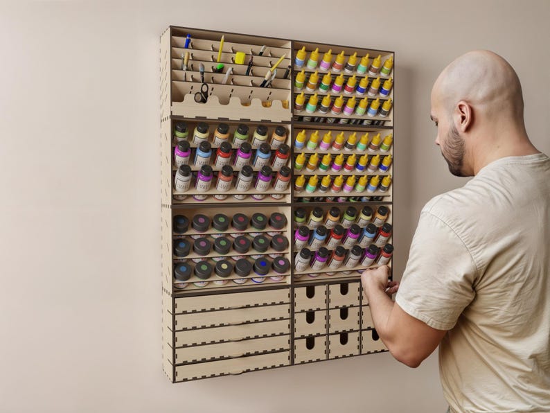 May include: A wooden wall-mounted organizer with multiple shelves and drawers for storing and displaying small bottles of paint. The organizer is made of light wood and has a natural finish.