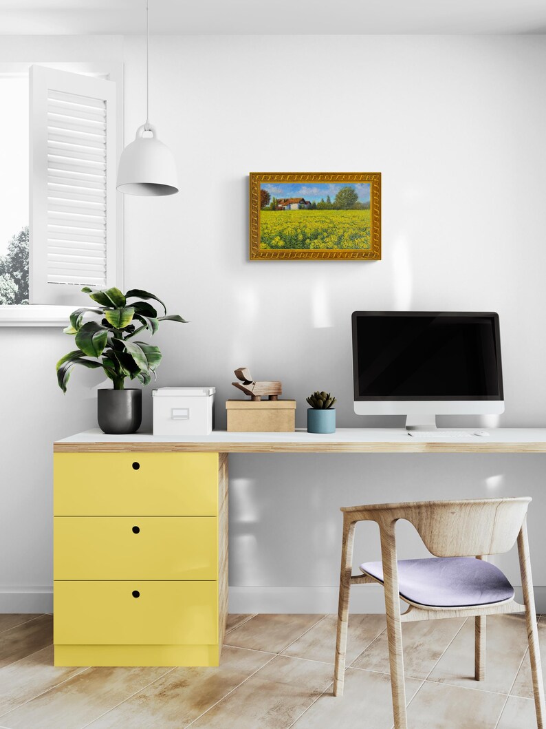 May include: A framed painting of a yellow field with a small house in the distance. The painting is hanging on a white wall above a desk with a computer, a chair, and a yellow drawer unit.