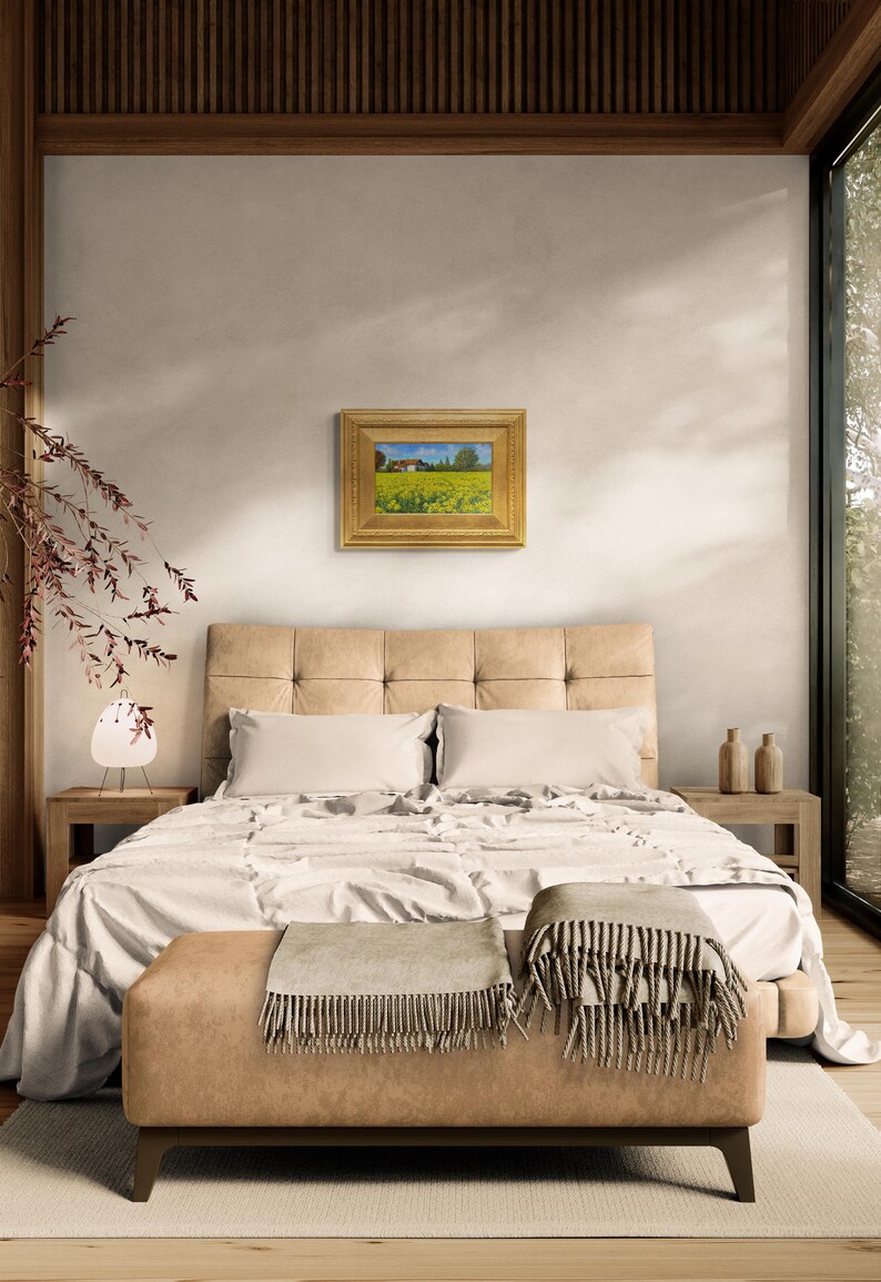 May include: A bedroom interior with a bed, a nightstand, and a framed landscape painting on the wall. The bed has a white duvet and two pillows. The nightstand has a lamp and a vase with flowers. The painting depicts a field of yellow flowers with a house in the background.
