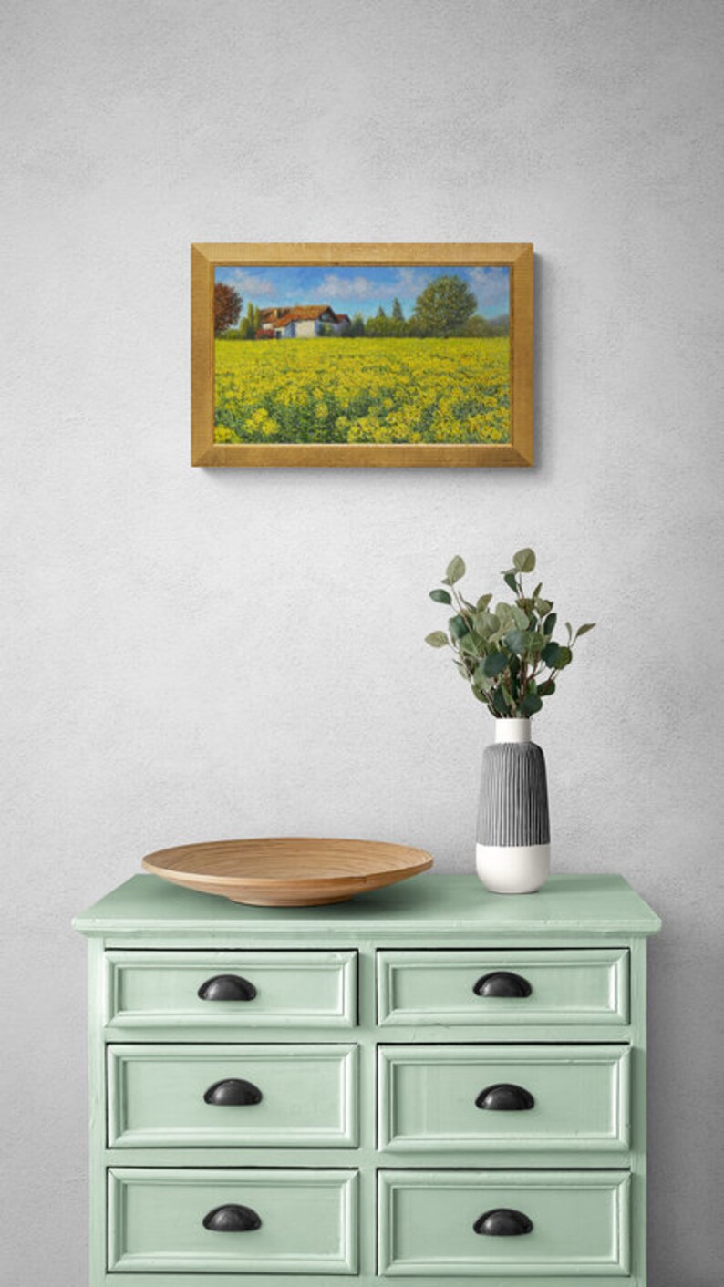 May include: A framed painting of a yellow field with a white farmhouse in the distance. The painting is hanging on a white wall above a light green dresser with six drawers.