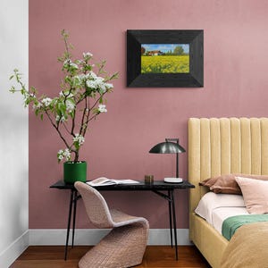 May include: A black framed picture of a field of yellow flowers with a house in the distance hangs on a pink wall. A black desk with a black lamp and a woven chair sits in front of the wall. A yellow headboard with a white bedspread and green throw blanket is in the background.