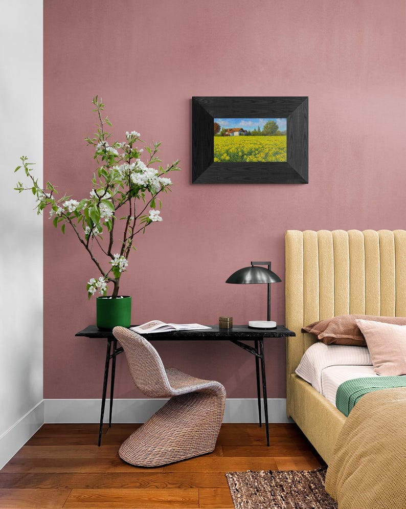 May include: A black framed picture of a field of yellow flowers with a house in the distance hangs on a pink wall. A black desk with a black lamp and a woven chair sits in front of the wall. A yellow headboard with a white bedspread and green throw blanket is in the background.