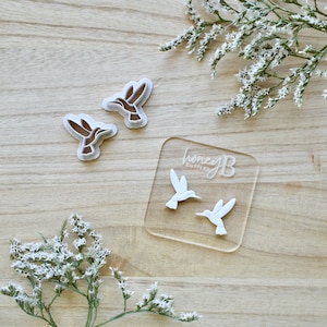 May include: Hummingbird-shaped cookie cutters in two styles. Two cutters have a brown hummingbird design with a white border. Two white hummingbird cutters are on a clear acrylic square with "honeyB cutters" printed on it. White flowers are in the background.