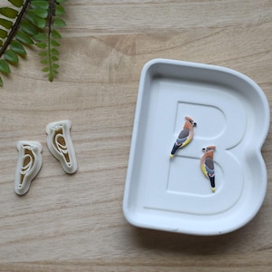 Könnte beinhalten: Ein weißes Keramiktablett in Form des Buchstabens "B" enthält zwei Vogelfiguren. Zwei Vogelförmige Ausstecher befinden sich links. Die Vögel sind braun, gelb und schwarz. Ein grüner Farnzweig befindet sich im Hintergrund.