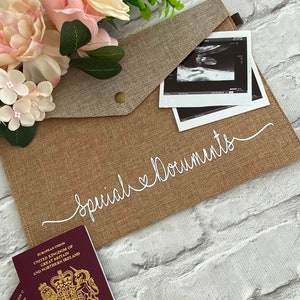 May include: A brown burlap envelope with a white inscription that reads "Special Documents". The envelope has a flap with a brass button closure. A passport and a black and white ultrasound image are visible inside the envelope.