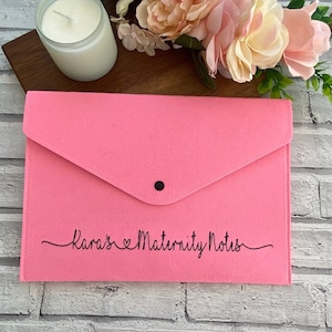 May include: A pink felt folder with a black button closure and the text "Kara's Maternity Notes" in black cursive lettering.
