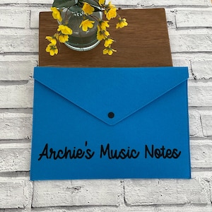 May include: A blue felt folder with a black button closure and the text "Archie's Music Notes" in black.