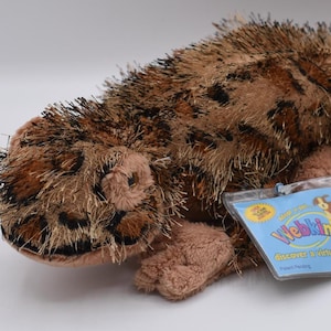 Peut inclure: Une peluche de gecko léopard avec des taches marron et noires, une longue queue et une étiquette « Webkinz ». Le jouet est fait d'une matière douce et floue, avec un ventre et des pattes brun clair.