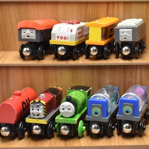 May include: Wooden toy train set featuring various colorful train cars with smiling faces. The set includes a red tanker car, a green engine, and cars with the text "FOG!" and a gray coal car. The trains are displayed on wooden shelves.