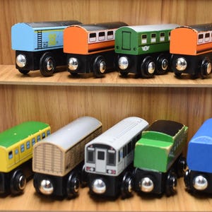Può includere: Vagoni di treno giocattolo in legno di vari colori, tra cui blu, arancione, verde e giallo. Ogni vagone ha un design unico ed è montato su ruote nere. I giocattoli sono esposti su una mensola di legno.