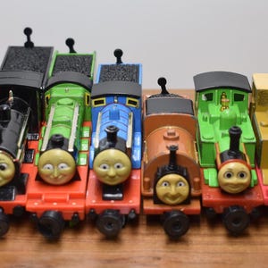 May include: A row of colourful toy train engines, featuring black, green, blue, brown, and yellow locomotives. Each train has a smiling face. They are displayed on a wooden surface. The engines are arranged in a line.