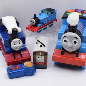 May include: A collection of Thomas the Tank Engine toys including two large blue and red train engines with white smoke stacks, one smaller blue train engine, a brown tram engine, and a blue handheld remote control with red and gold buttons.