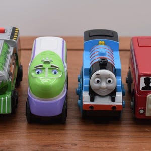 May include: Four toy vehicles: a green and grey train, a green and purple train, a blue train with a face, and a red bus with a face. The toys are arranged in a row on a wooden surface.