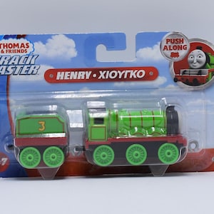 May include: Thomas & Friends Track Master Henry toy train with a green and red engine and a green and red caboose. The train is on a white background with the words "Track Master" and "Henry - Xioytko" printed on the packaging. The packaging also has a picture of Thomas the Tank Engine on it.