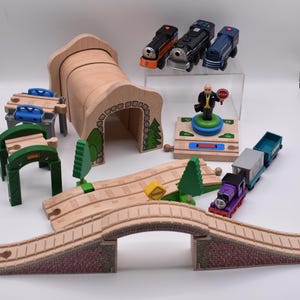May include: A wooden toy train set featuring a tunnel, bridge, tracks, and various train cars. The set includes a stop sign figure and several colorful train engines. The bridge has a brick design.