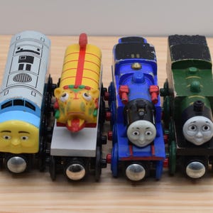 May include: A set of four toy trains including a light blue and yellow diesel engine, a yellow dragon-themed cargo car, a blue steam engine with a face, and a green steam engine with a face, all featuring black wheels and metal connectors.