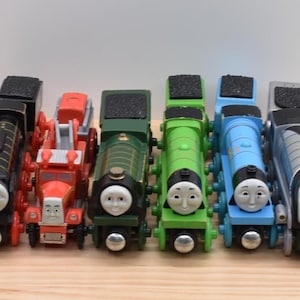 May include: A collection of six toy train engines and a fire engine vehicle lined up side by side. The set includes a black engine, a red fire truck, a dark green engine, a bright green engine, a blue engine, and a silver streamlined engine.