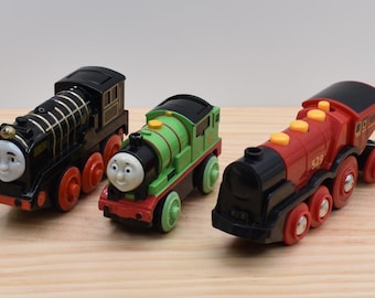 May include: Three toy trains featuring character faces. The first is a black locomotive with gold stripes and red wheels. The second is a green locomotive with a black roof and light green wheels. The third is a red locomotive with yellow accents.