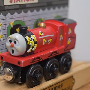 May include: A red wooden toy train engine with a black face and yellow and black bees painted on the sides. The engine is on a wooden track and has the word "James" written on the track.