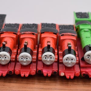 May include: A collection of toy train engines in various colors. The engines include a black engine, several red engines, and two green engines. Each engine has a smiling face and is designed to resemble a classic steam train.
