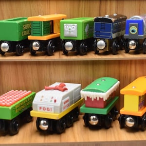 May include: A selection of brightly coloured wooden toy train carriages displayed on wooden shelves. The carriages feature various designs, including green, yellow, and blue, with details like a face, text, and decorative elements. The carriages are on black wheels.