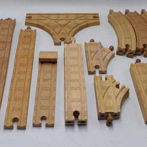May include: A collection of wooden toy train tracks in various shapes and sizes. The set includes straight, curved, and switch tracks, as well as a bridge piece. The tracks are made of light-colored wood and are designed for a toy train set.