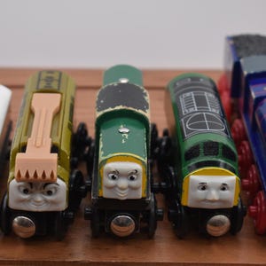 Può includere: Cinque trenini giocattolo in legno di vari colori, tra cui bianco, verde oliva, verde scuro e blu. Ogni trenino ha un volto e un design unici, con ruote rosse e nere. I trenini sono disposti in fila su una superficie di legno.