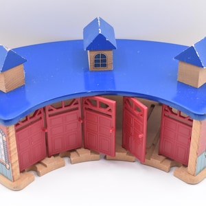 Könnte beinhalten: Ein hölzerner Spielzeugbahnhof mit blauem Dach und roten Türen. Der Bahnhof hat ein gebogenes Design mit drei kleinen Strukturen mit blauem Dach oben. Die Türen sind geöffnet und geben den Blick ins Innere frei.